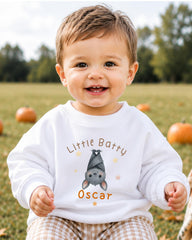 A smiling baby boy wearing a white sweatshirt with a bat design and the name "Oscar" printed on it, sitting in a field with pumpkins in the background.