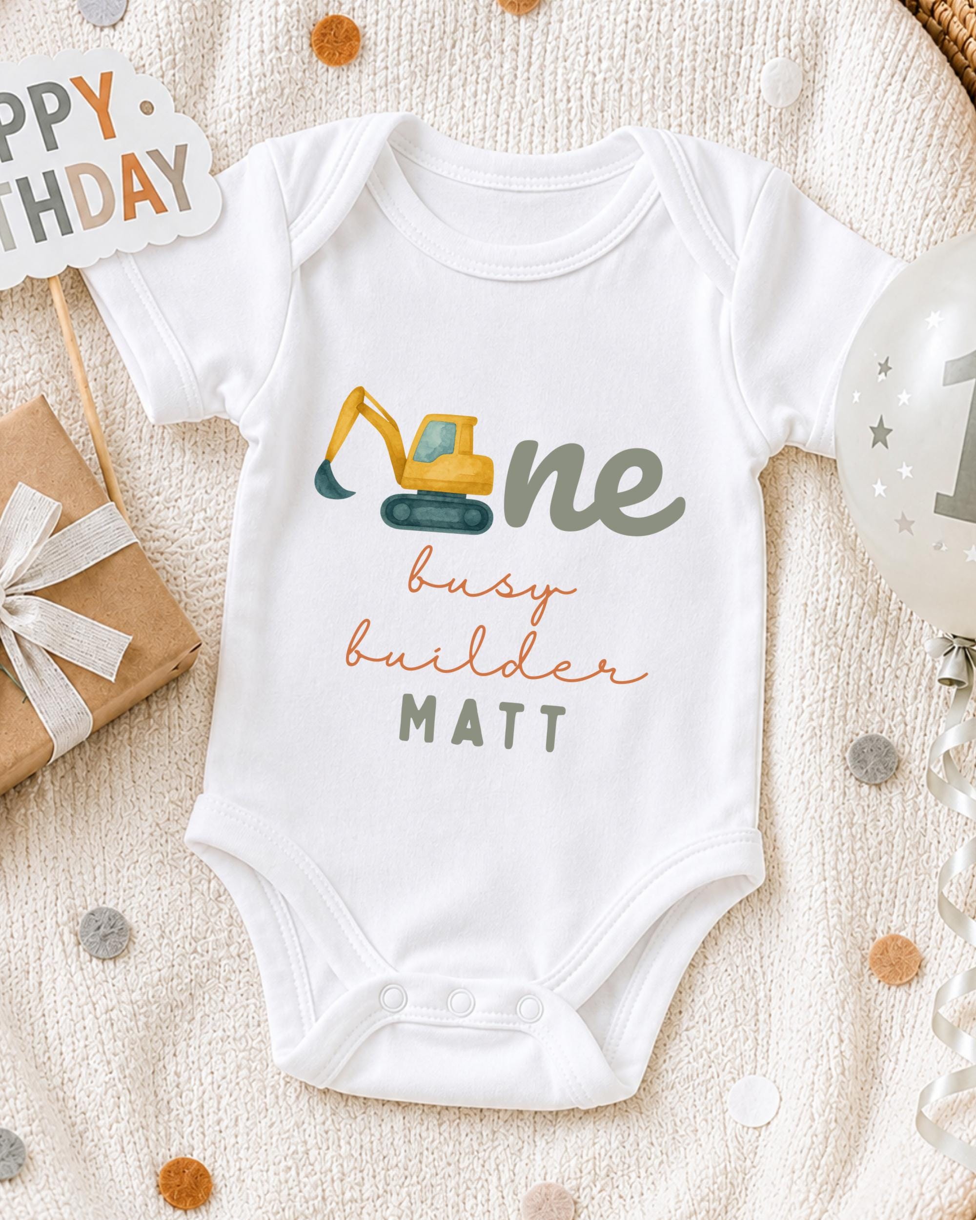 A white onesie with a construction vehicle design, surrounded by party decorations and a birthday sign.