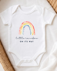 A white baby onesie with a rainbow graphic and the text "little rainbow on its way" printed on it.
