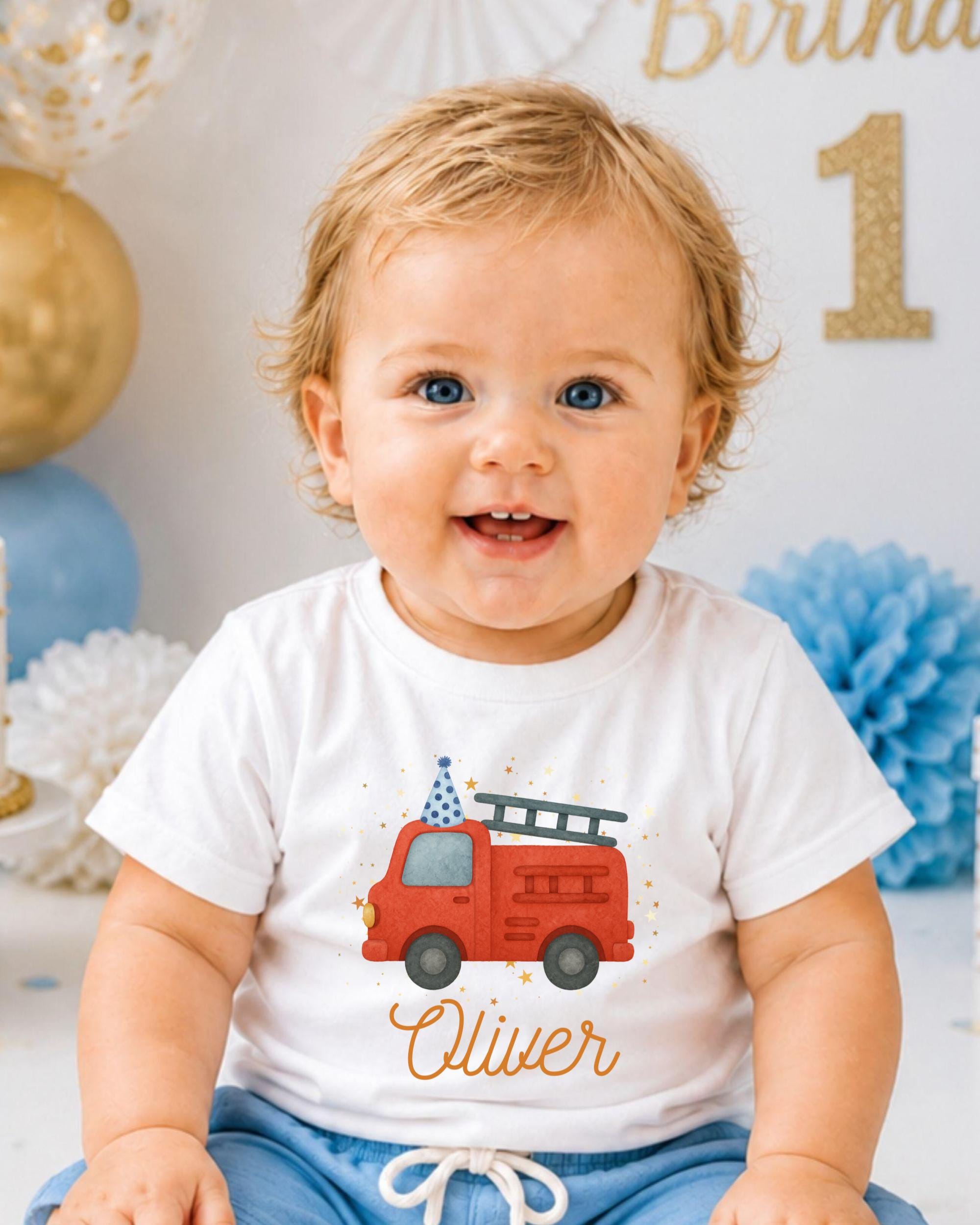 A smiling baby boy wearing a white t-shirt with a red fire truck graphic and the name "Olivier" printed on it, sitting in front of a birthday party backdrop.