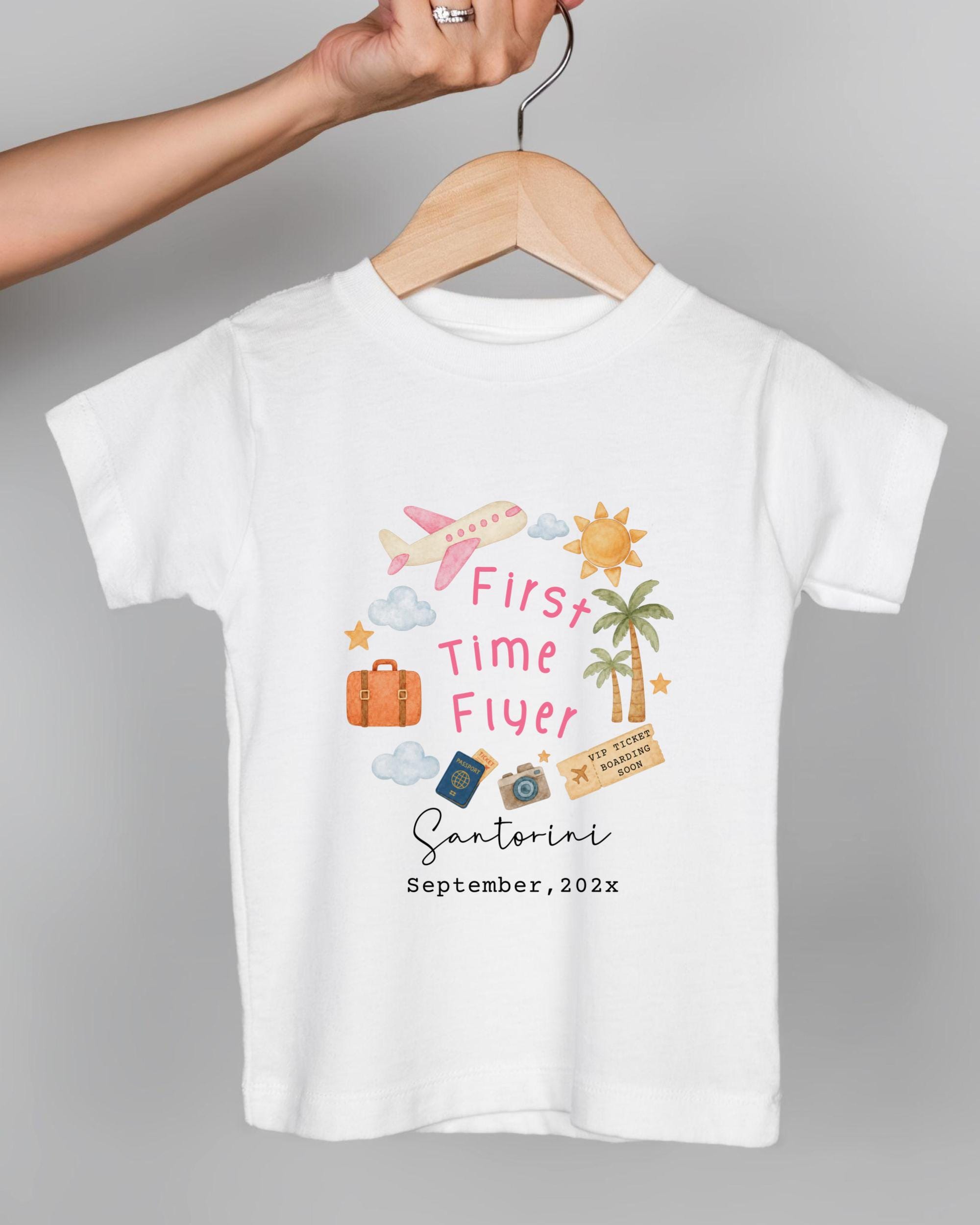 A white t-shirt with a graphic design featuring various travel-related icons and text, including a plane, a suitcase, a beach, and a palm tree.