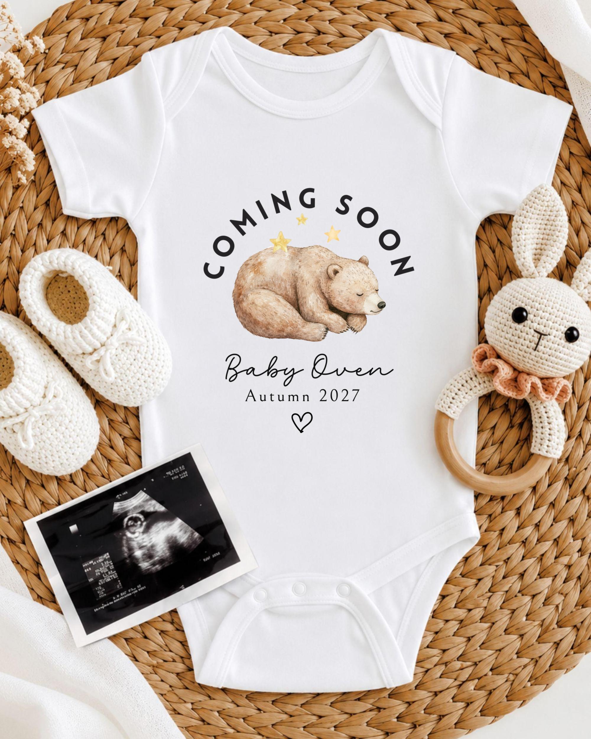 A baby announcement t-shirt with a bear image and the text "Coming soon Baby Queen Autumn 2027" is displayed alongside a crocheted rabbit toy and a baby's foot.