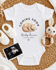 A baby announcement t-shirt with a bear image and the text "Coming soon Baby Queen Autumn 2027" is displayed alongside a crocheted rabbit toy and a baby's foot.