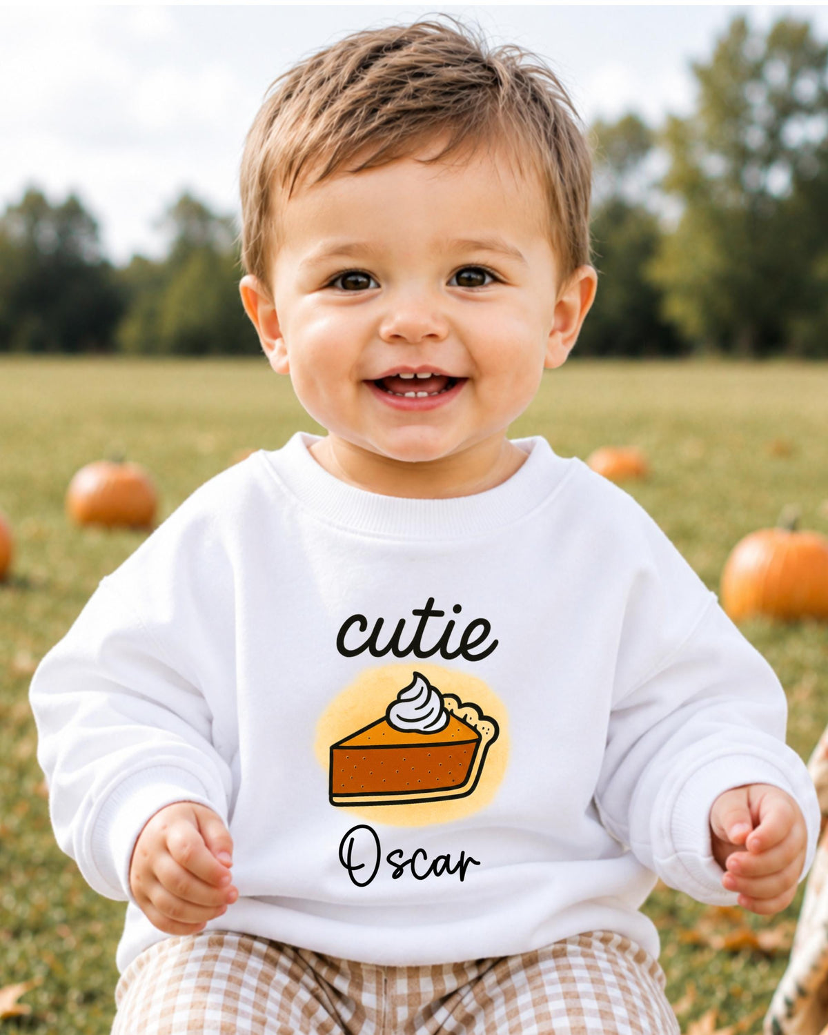 A smiling baby boy wearing a white sweater with a pumpkin and whipped cream design, sitting in a field with pumpkins in the background.