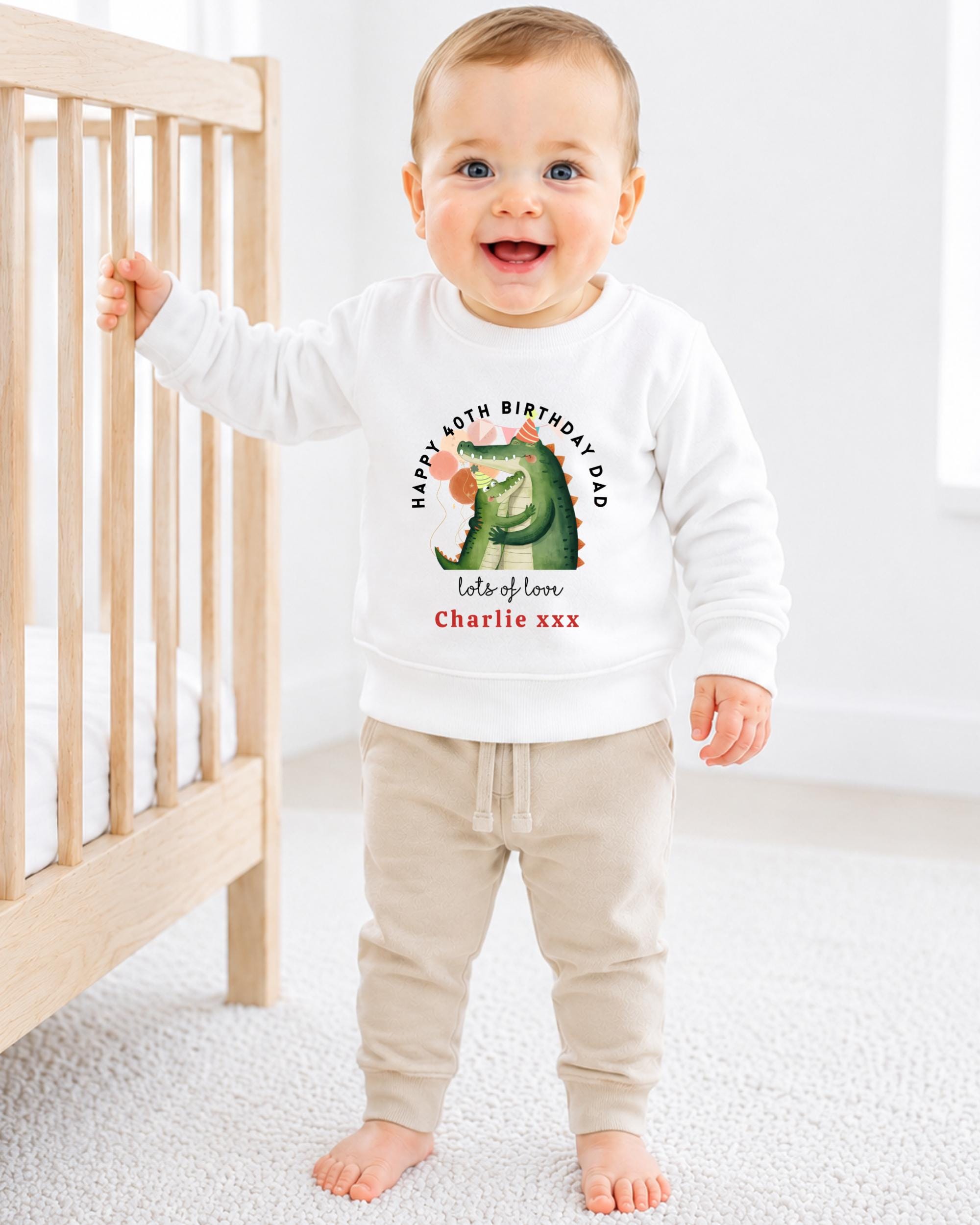 A smiling baby boy wearing a white sweatshirt with a dinosaur graphic and text on it, standing next to a wooden crib.