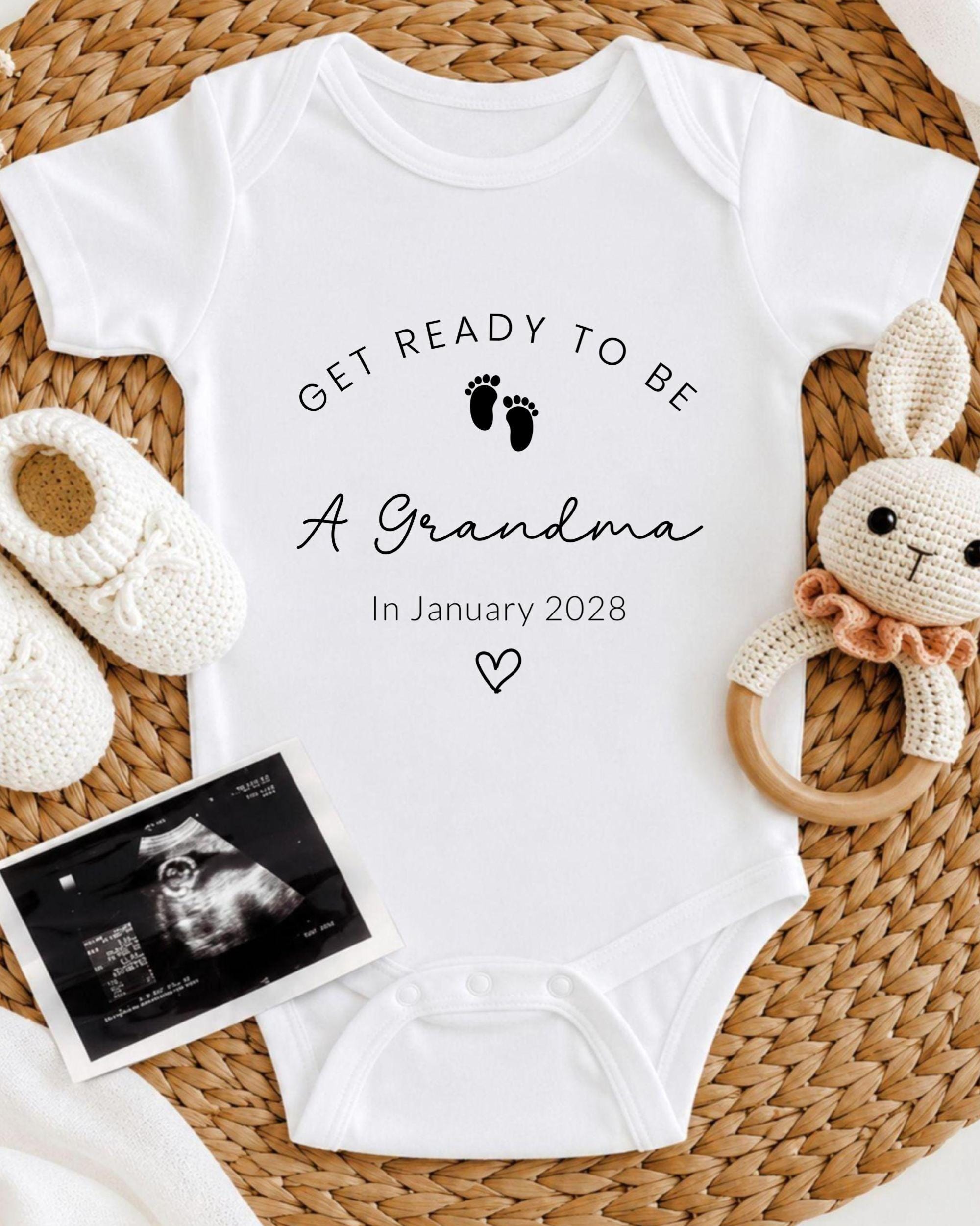 A white baby onesie with the text "GET READY TO BE A GRANDMA In January 2028" printed on it, along with a pair of white crocheted shoes and a stuffed rabbit.