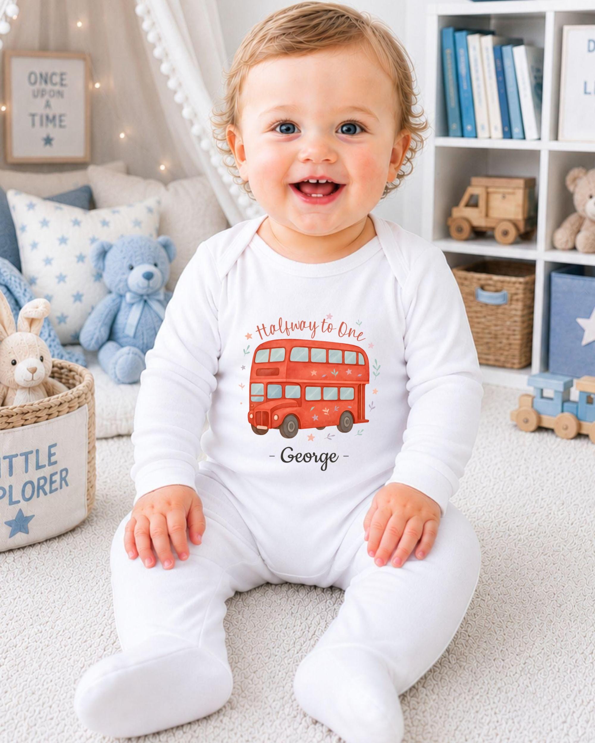 A smiling baby boy wearing a white onesie with a red double-decker bus design sits on the floor in a nursery setting.
