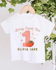 A white t-shirt with a pink number one and strawberries printed on it, hanging on a wooden hanger.