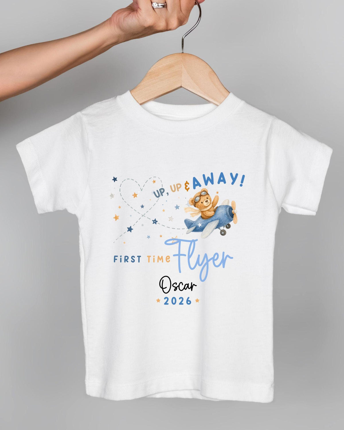 A white t-shirt with a graphic design featuring a cartoon character flying in a plane and the text "UP, UP & AWAY! FIRST TIME FLYER Oscar 2026".