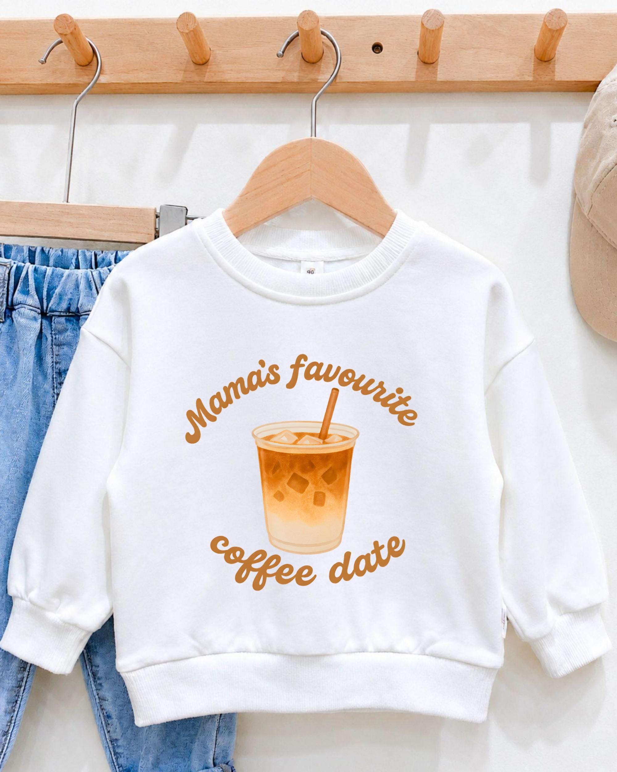 A white sweatshirt with a graphic of a coffee cup and the text "Mama's favourite coffee date" printed on it, hanging on a wooden hanger.