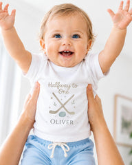A smiling baby wearing a white t-shirt with the text "Halfway to One - Oliver" and a graphic of a hockey stick and puck.