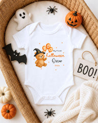 A white onesie with a teddy bear and a witch hat, along with Halloween decorations such as a pumpkin and a ghost, are arranged on a bed.