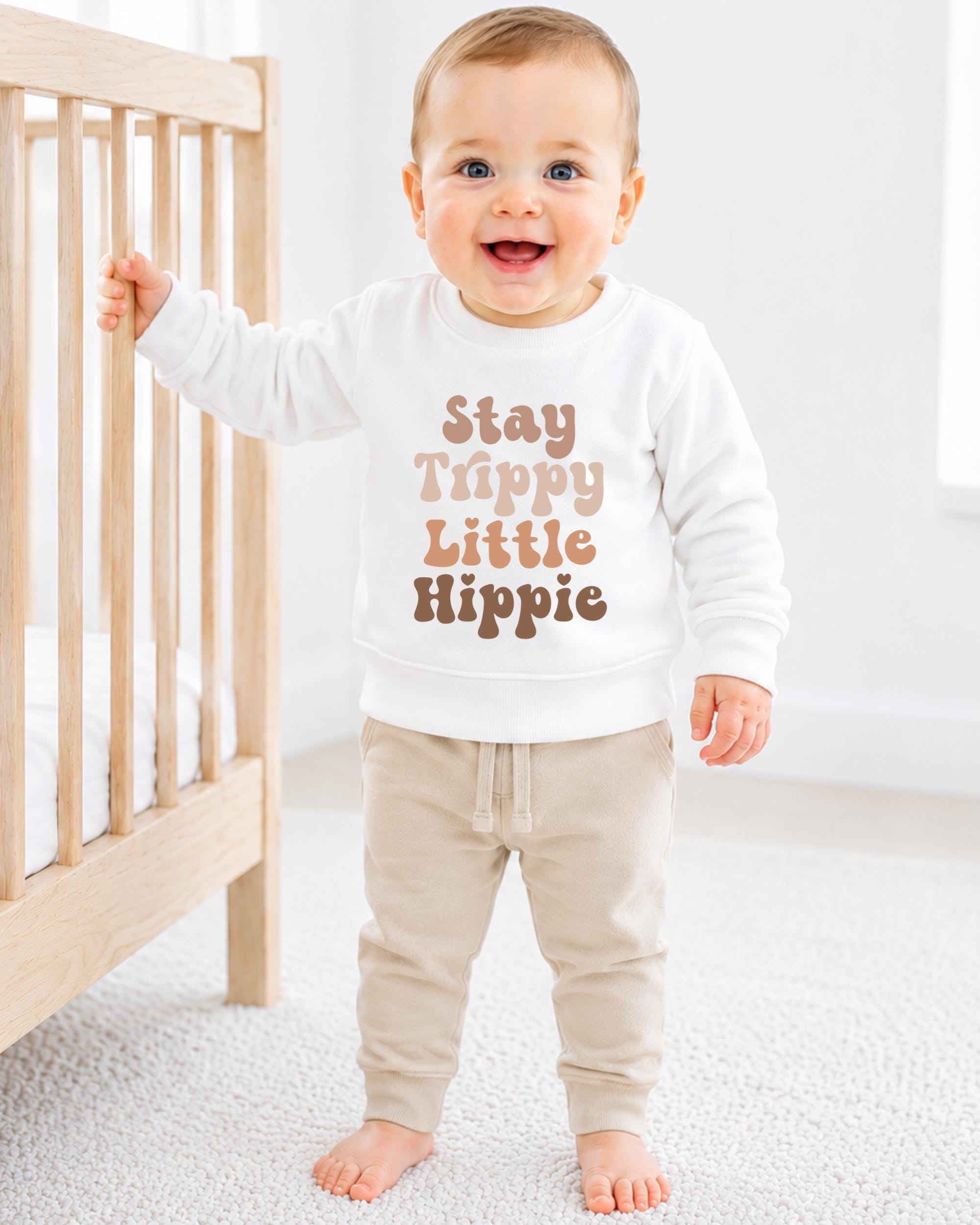 A smiling baby boy wearing a white sweatshirt with the text "Stay Trippy Little Hippie" printed on it, standing next to a wooden crib.