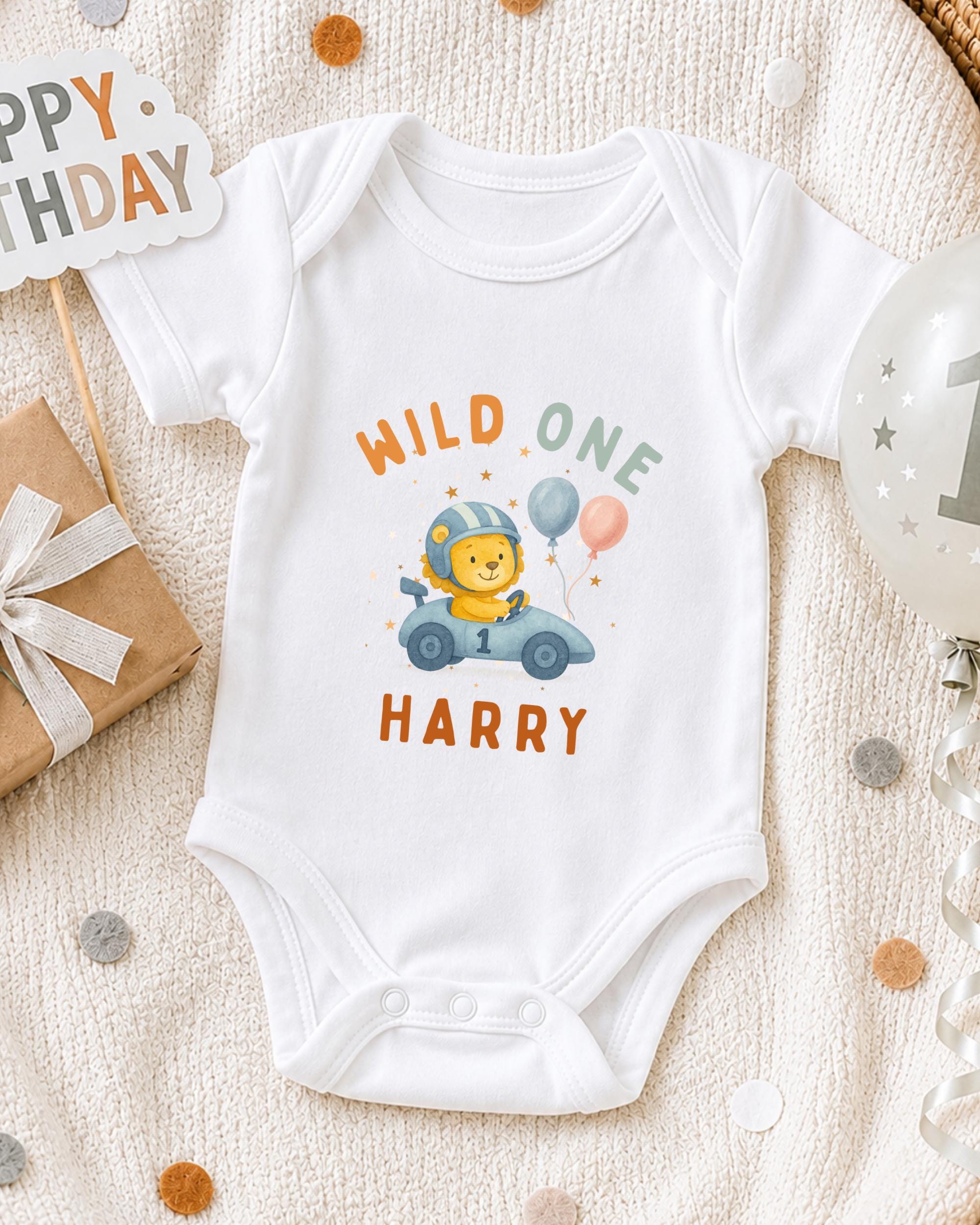 A white onesie with a cartoon lion and the text "WILD ONE HARRY" printed on it, along with a birthday sign and balloons, suggesting a celebration for a baby's first birthday.