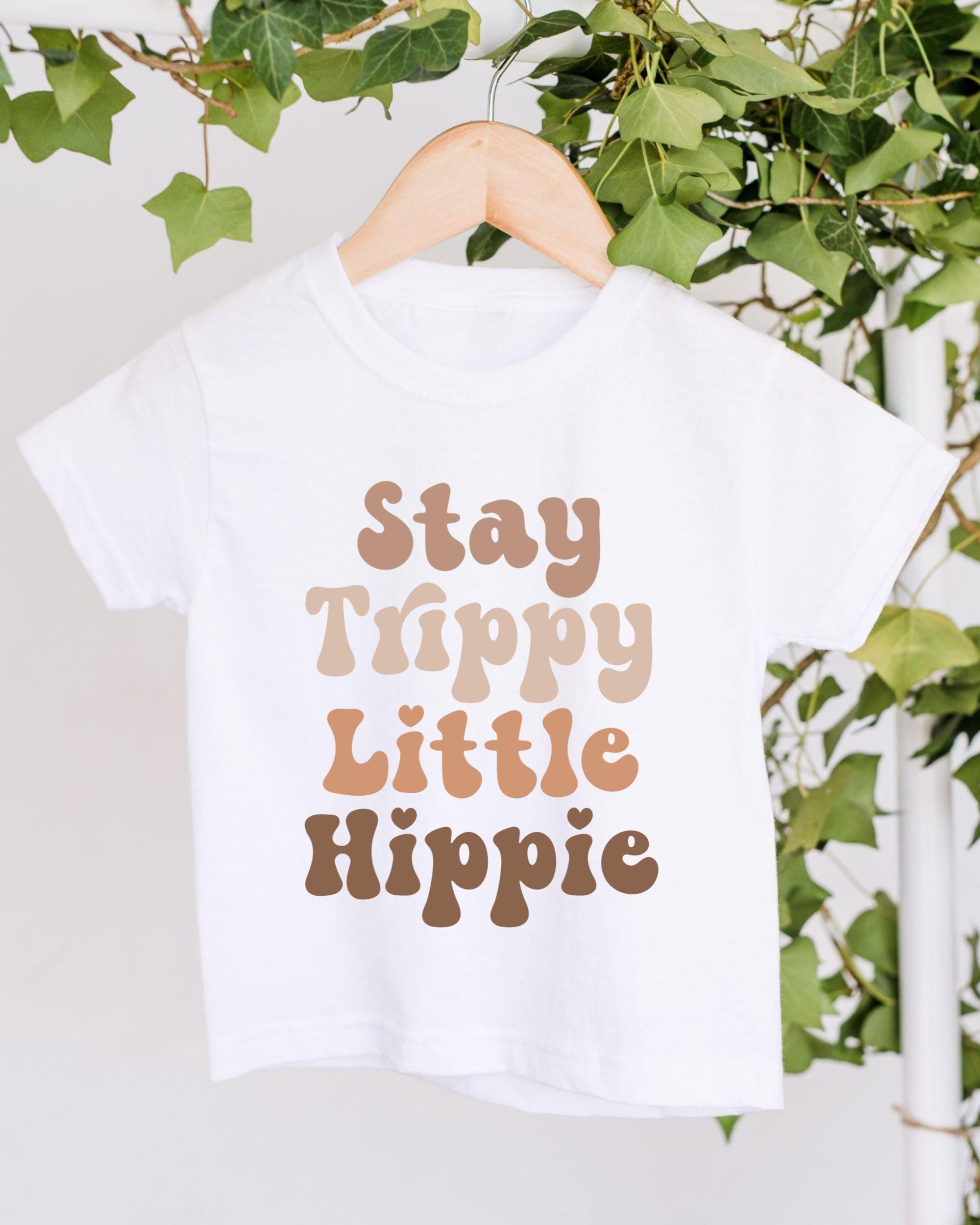 A white t-shirt with the text "Stay Trippy Little Hippie" printed on it, hanging on a wooden hanger.
