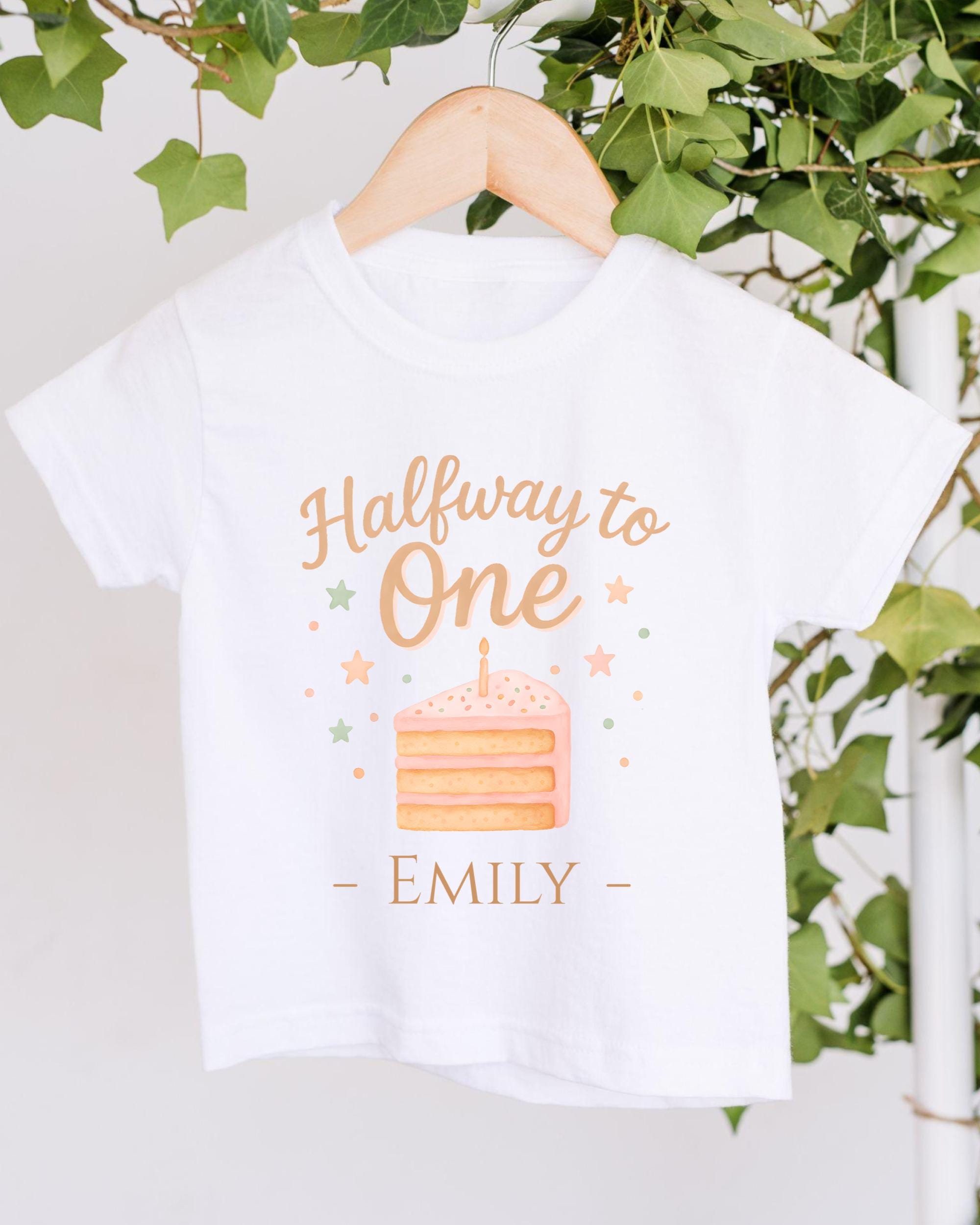 A white t-shirt with a graphic design featuring a slice of cake and stars, along with the text "Halfway to One - Emily" printed on it.