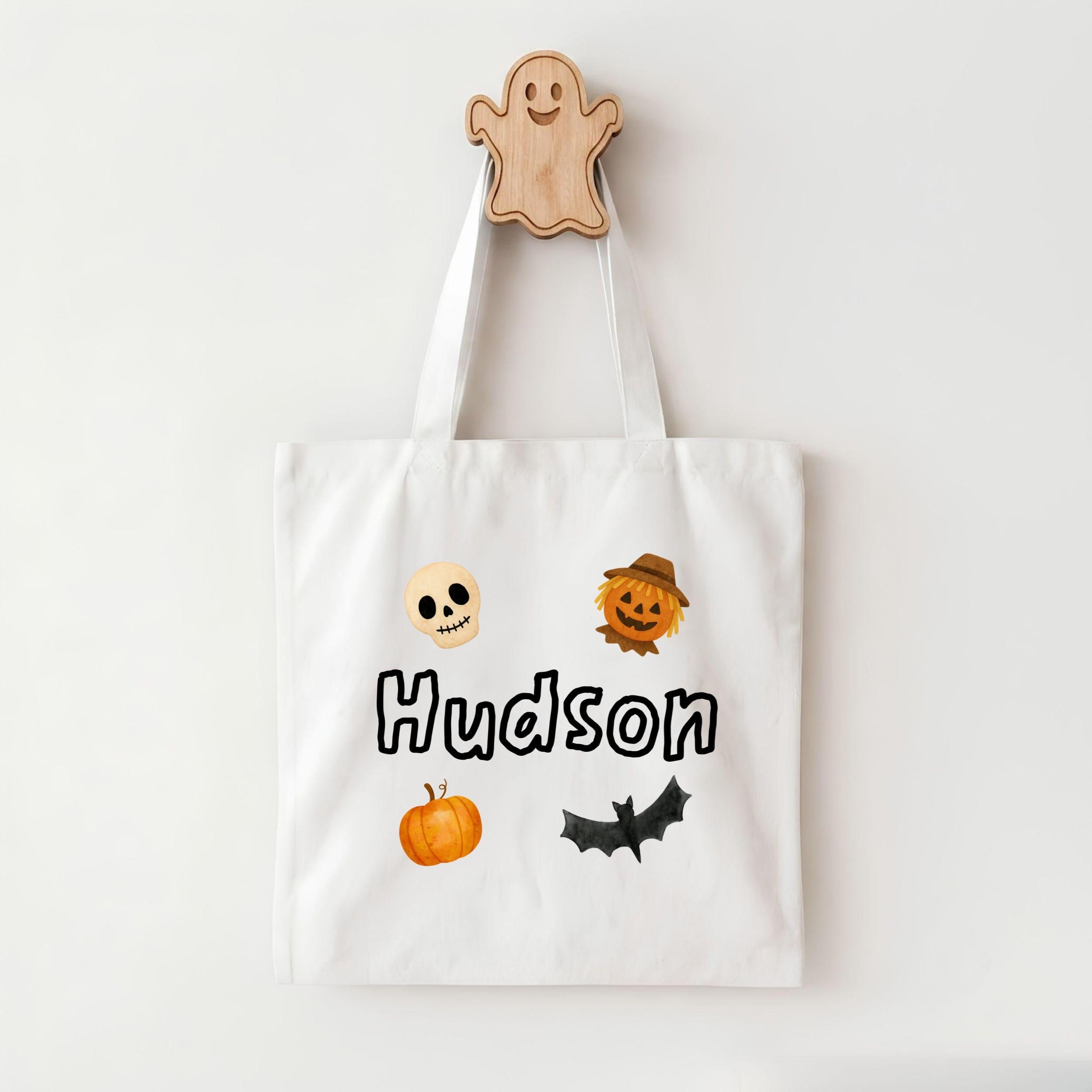 A white tote bag with a name printed on it, featuring a ghost, pumpkin, skeleton, scarecrow, and bat.