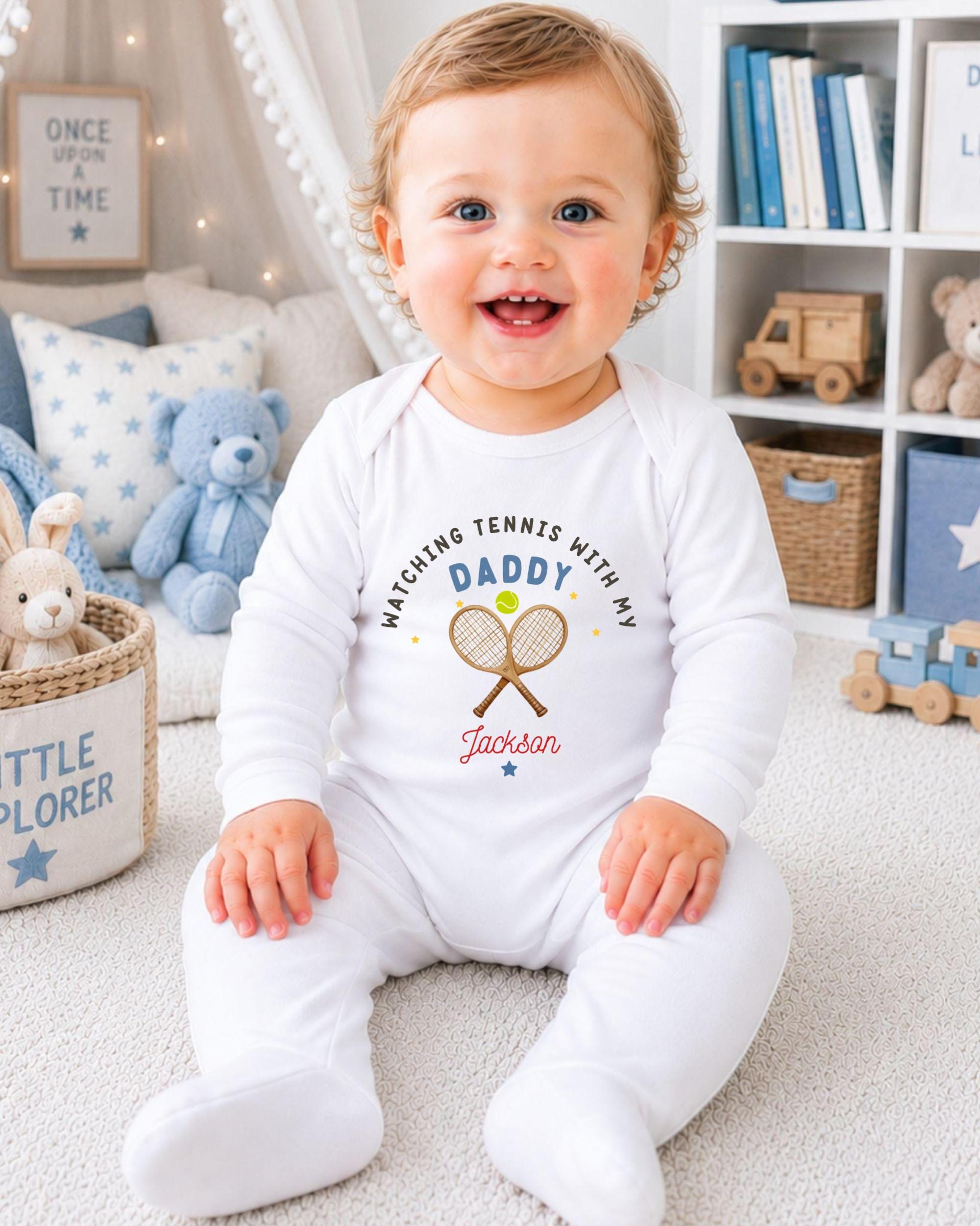 A smiling baby boy wearing a white onesie with a tennis racket and the text "Matching Tennis with my Daddy Jackson" is sitting on the floor in a nursery setting.