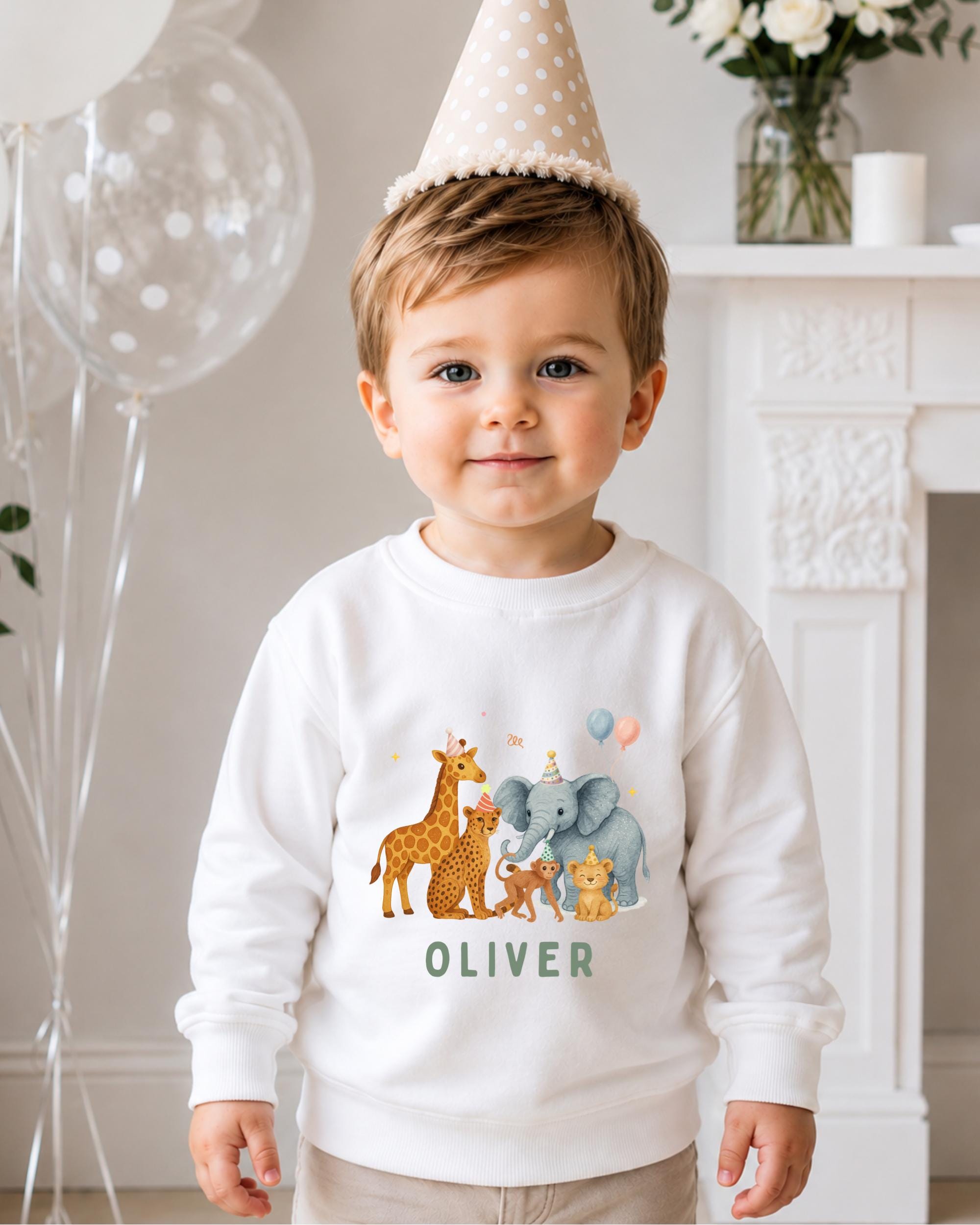 A young boy wearing a white sweater with a cartoon illustration of various animals, including giraffes, elephants, and a monkey, standing in front of a fireplace. The boy is wearing a birthday hat and has a name tag that says "OLIVER".