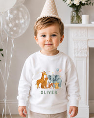 A young boy wearing a white sweater with a cartoon illustration of various animals, including giraffes, elephants, and a monkey, standing in front of a fireplace. The boy is wearing a birthday hat and has a name tag that says "OLIVER".