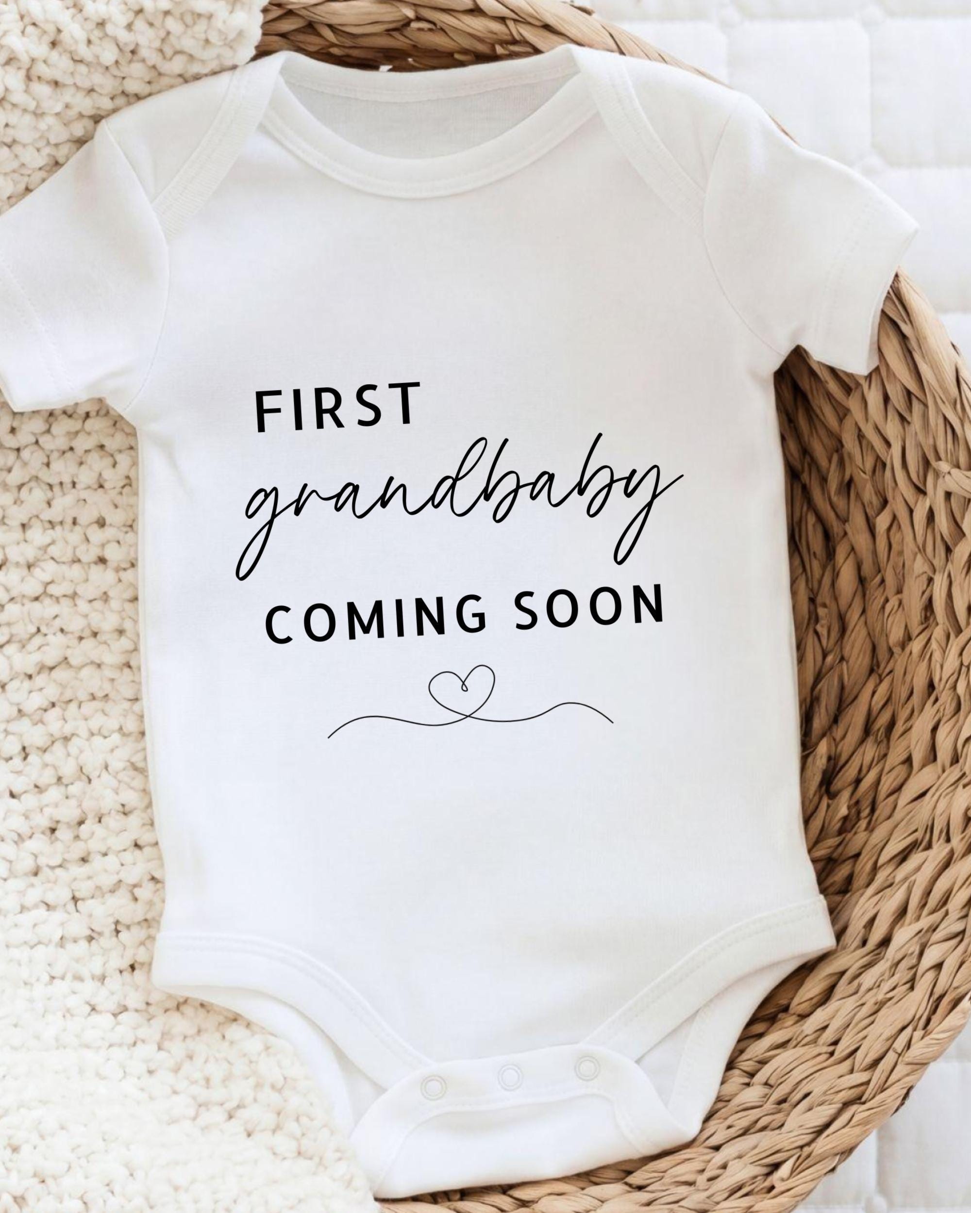 A white baby onesie with the text "FIRST GRANDBABY COMING SOON" printed on it, placed on a woven basket.