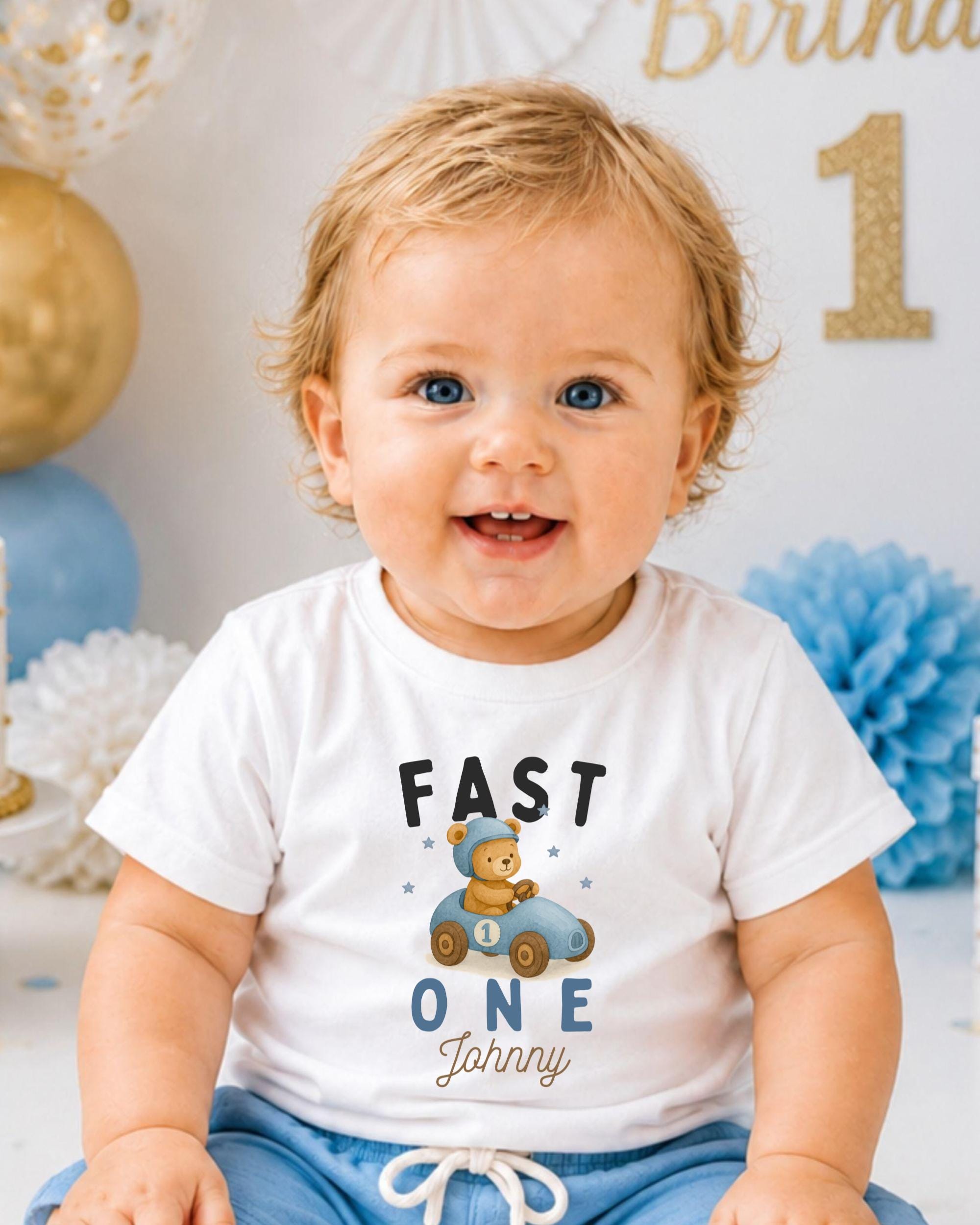 A smiling baby boy wearing a white t-shirt with a teddy bear and the text "Fast One Janna" printed on it.