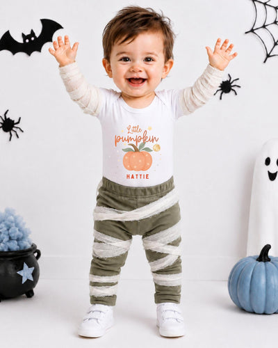 A smiling baby boy wearing a white t-shirt with a pumpkin graphic and striped pants, standing in front of Halloween decorations.
