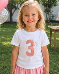 A young girl with blonde hair is smiling and wearing a white t-shirt with the number 3 and the name "Hannah" printed on it. She is also wearing a pink skirt.