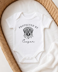 A white baby onesie with a graphic of a dog's head and the text "Protected by Cooper" printed on it.