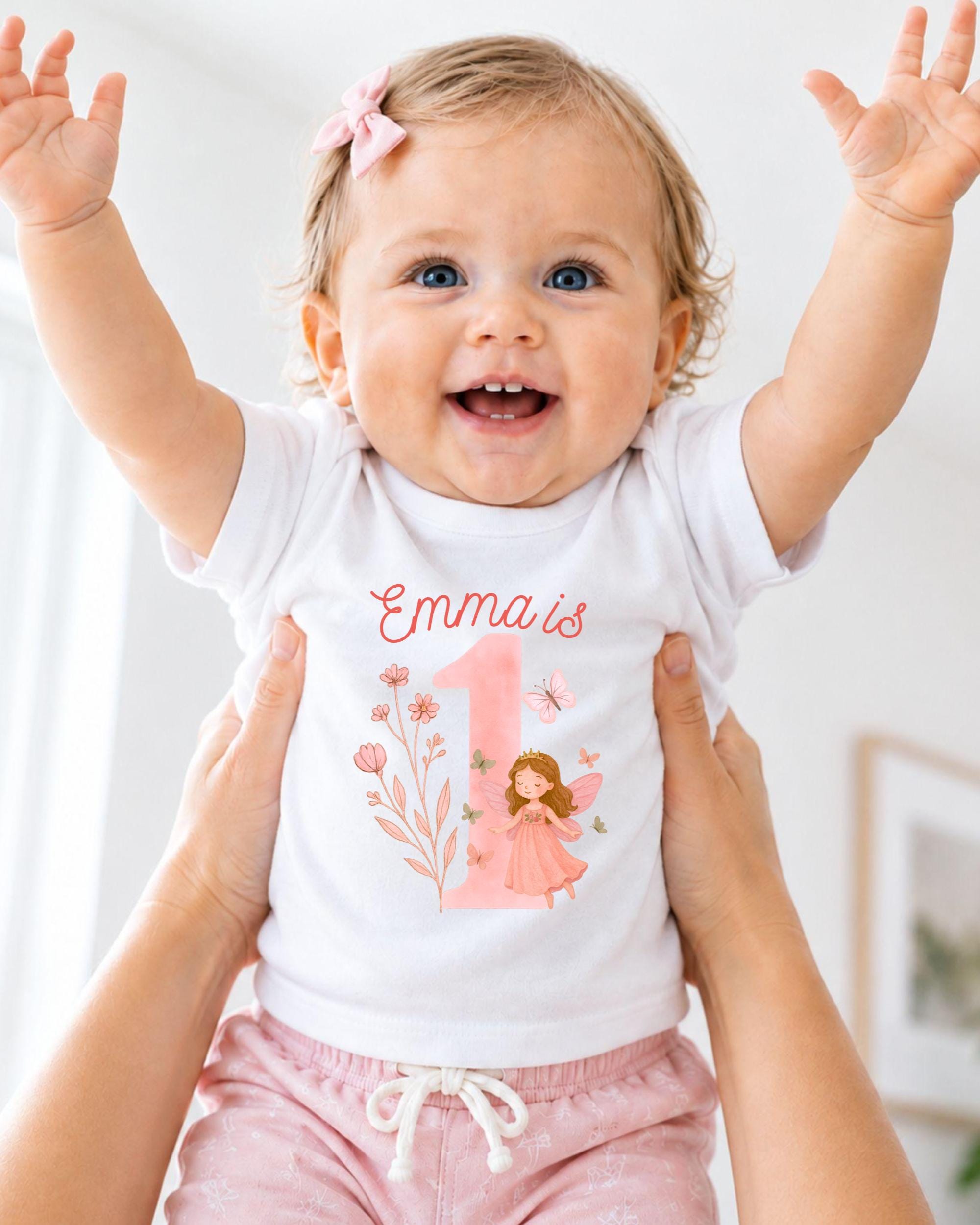 A smiling baby girl wearing a white t-shirt with a fairy design and the name "Emma" printed on it, being held up by someone's arms.