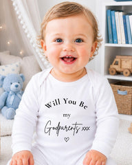 A smiling baby wearing a white onesie with text that says "Will You Be my Godparents xxx"