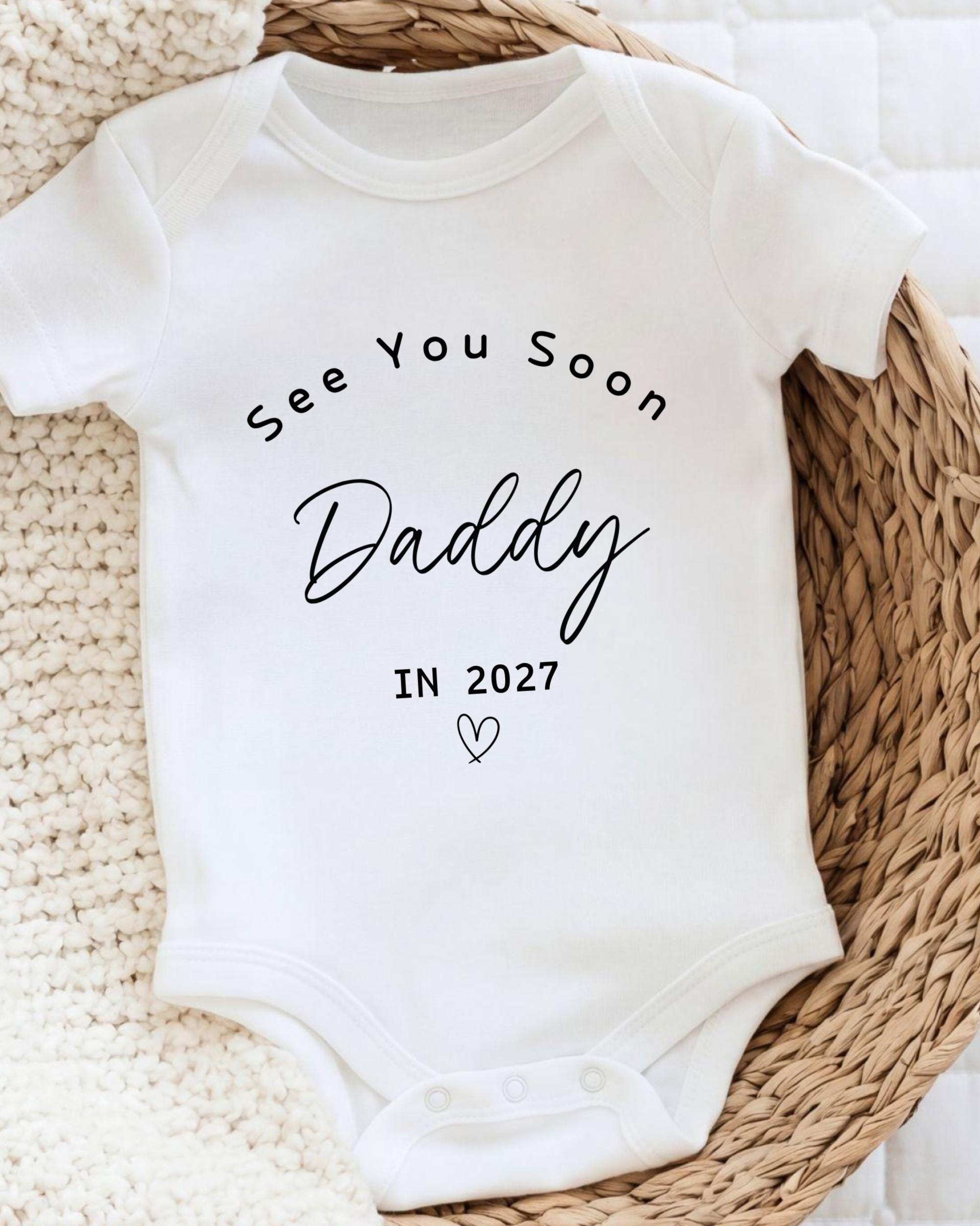 A white baby onesie with the text "See You Soon Daddy in 2027" printed on it.