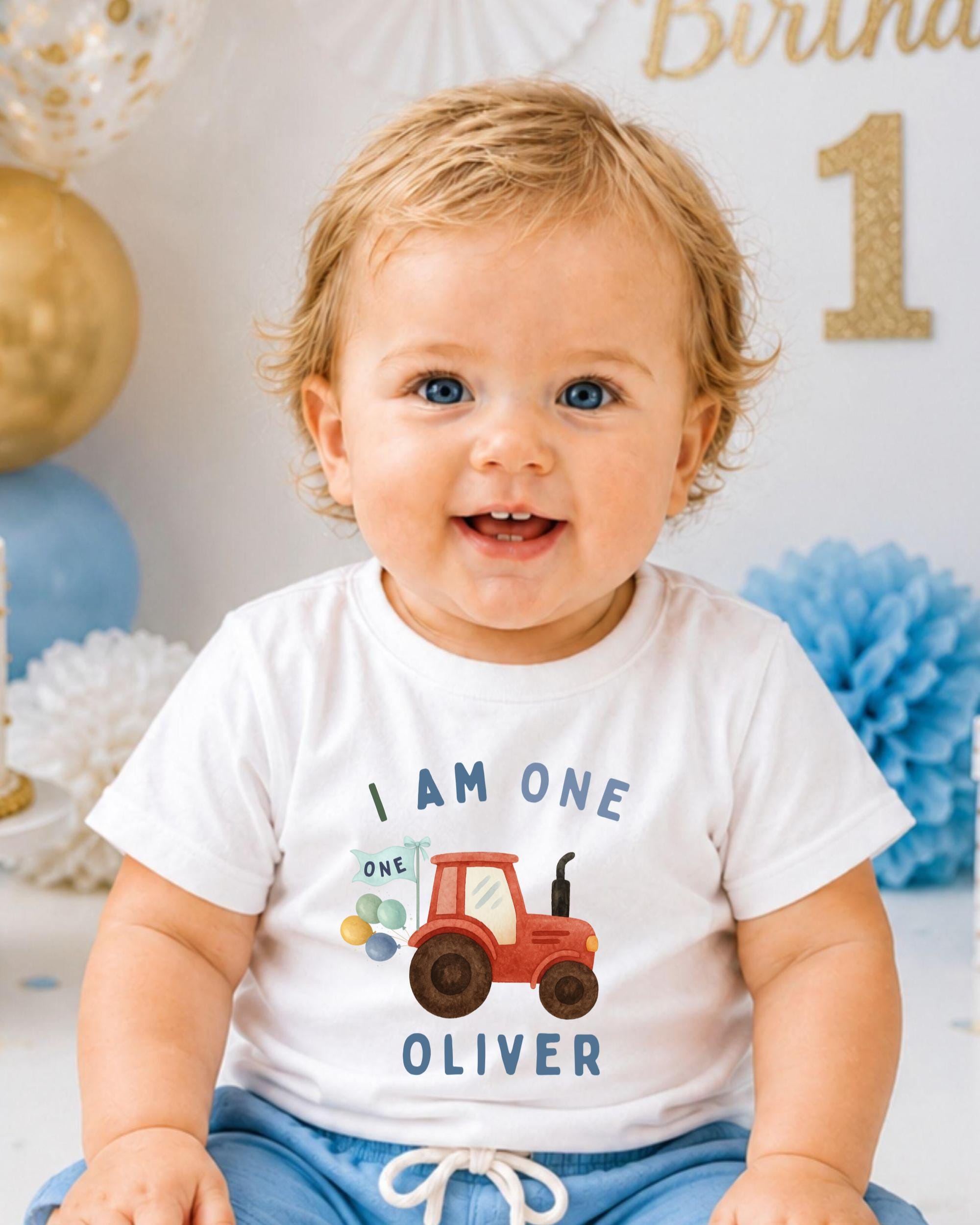 A smiling baby boy wearing a white t-shirt with a tractor graphic and the text "I AM ONE OLIVER" printed on it.