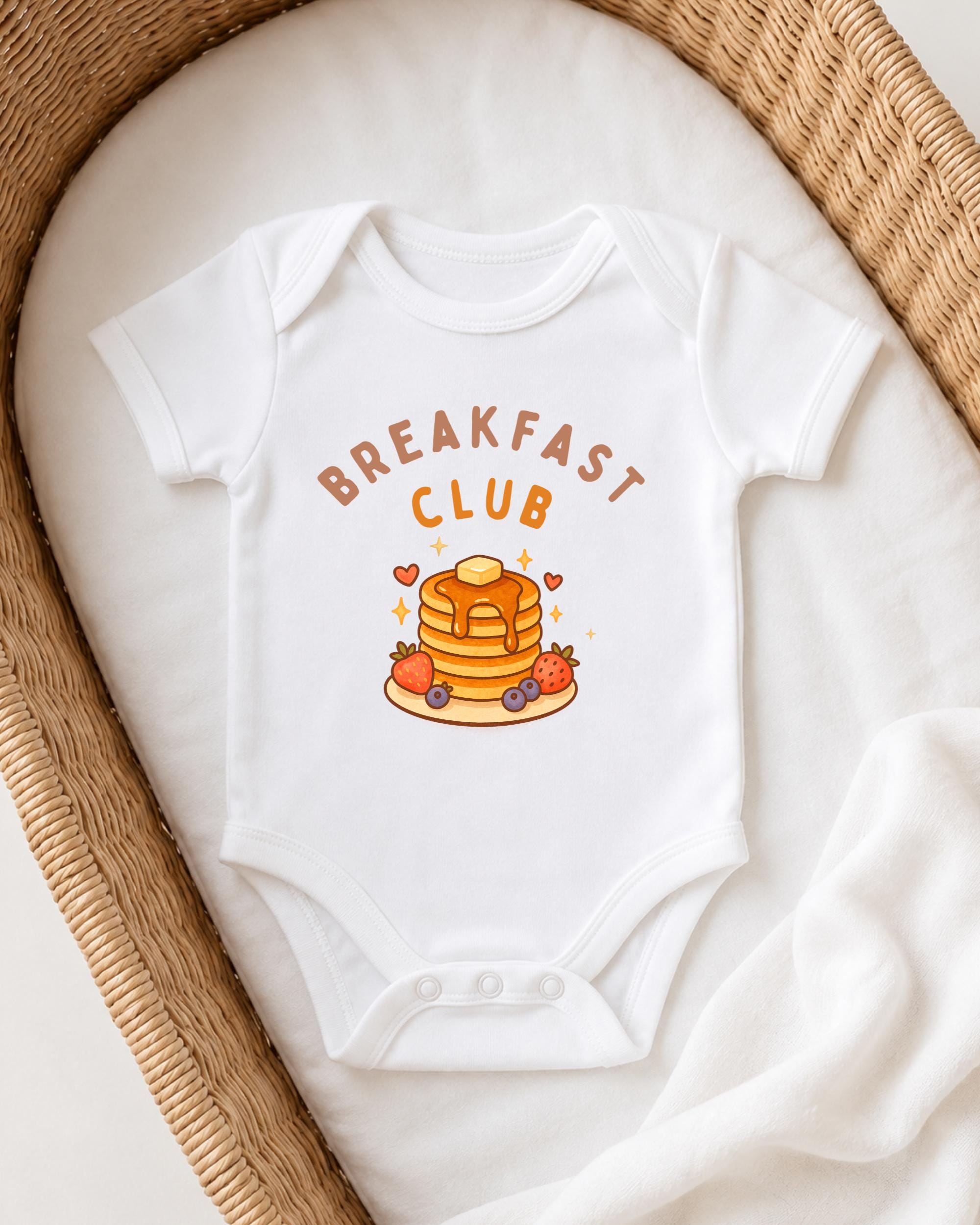 A white onesie with the text "BREAKFAST CLUB" printed on it, featuring a stack of pancakes and various breakfast-themed illustrations.