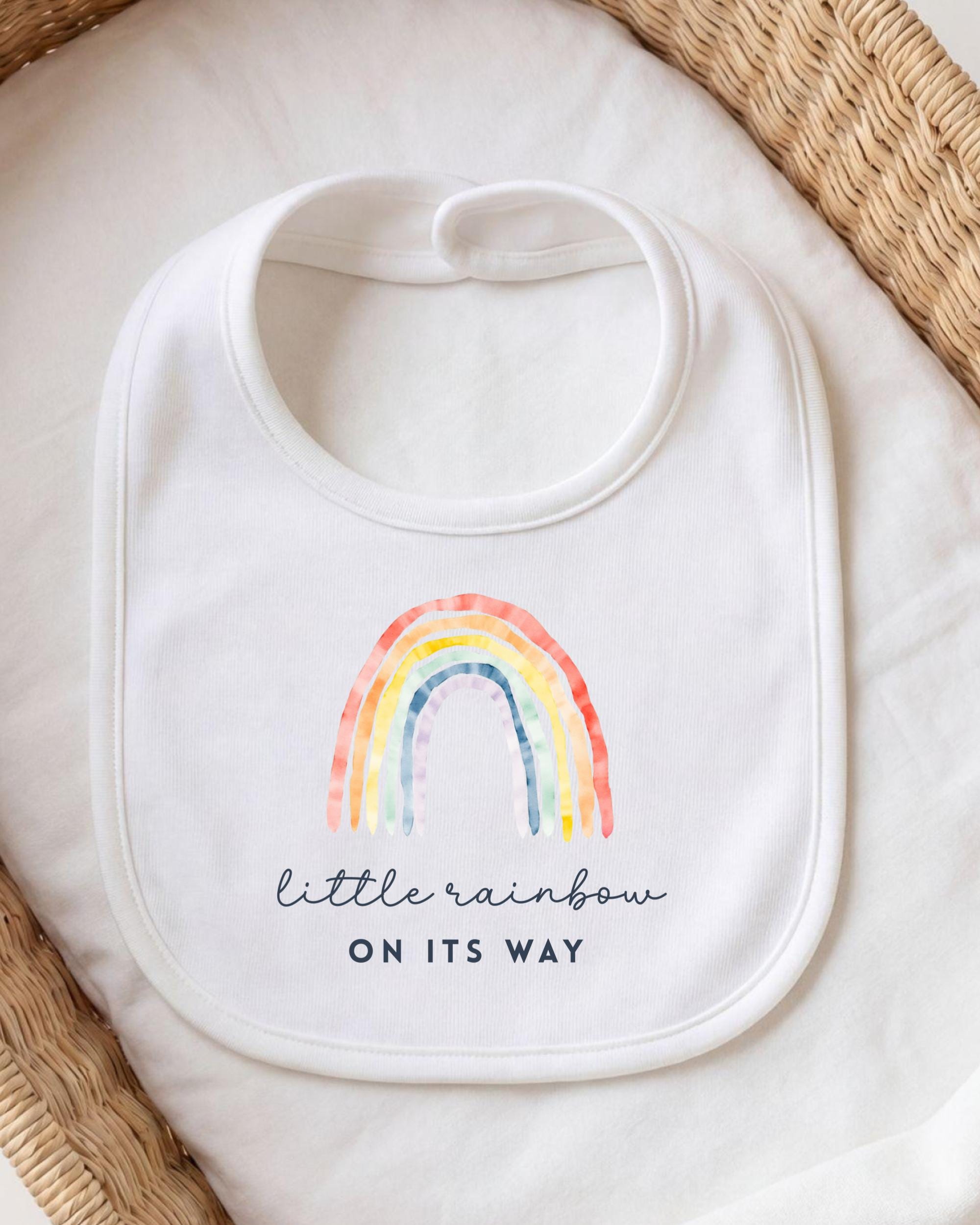 A white bib with a rainbow graphic and the text "little rainbow on its way" printed on it.