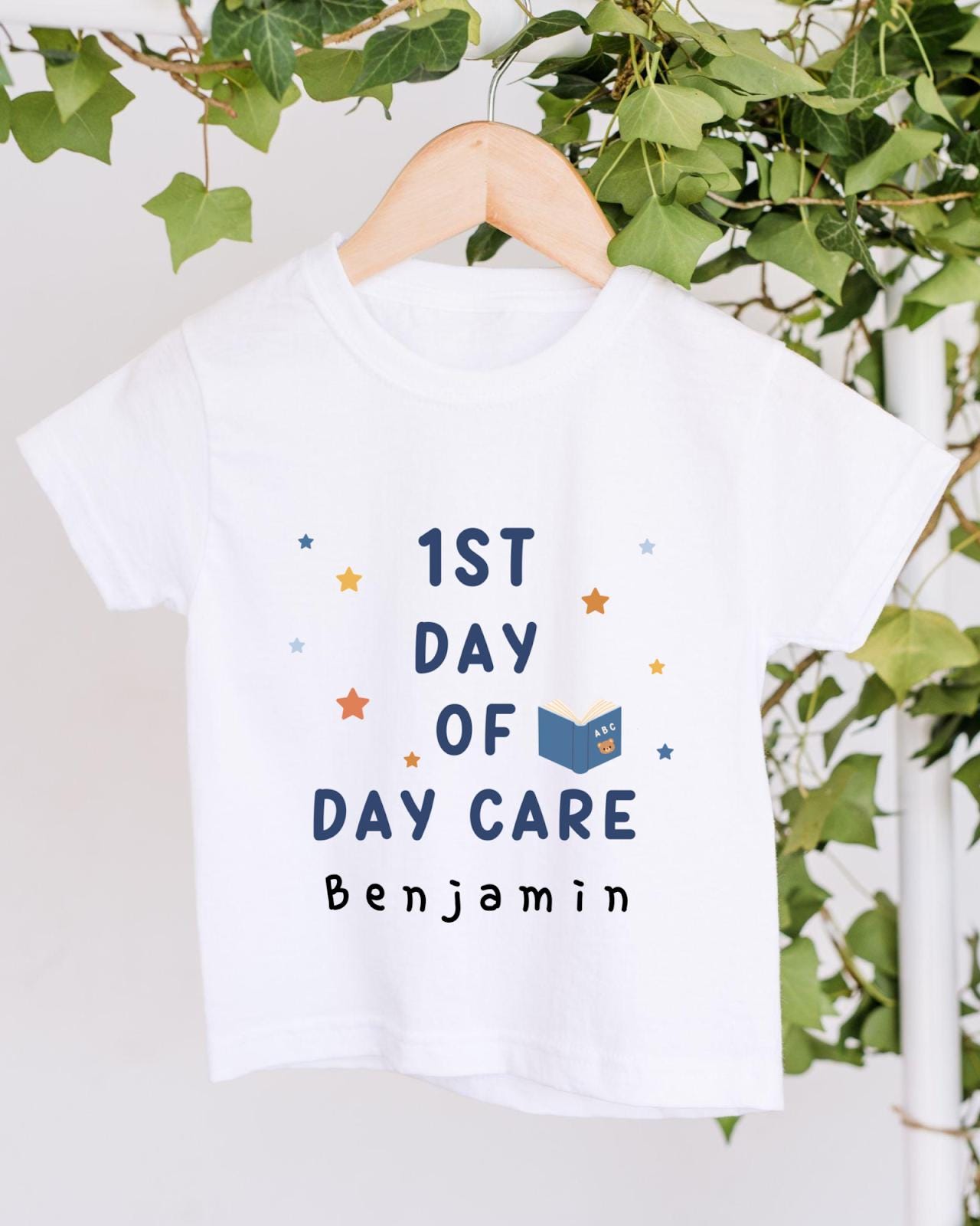 A white t-shirt with the text "1st DAY OF DAY CARE Benjamin" printed on it, along with stars and a book icon.