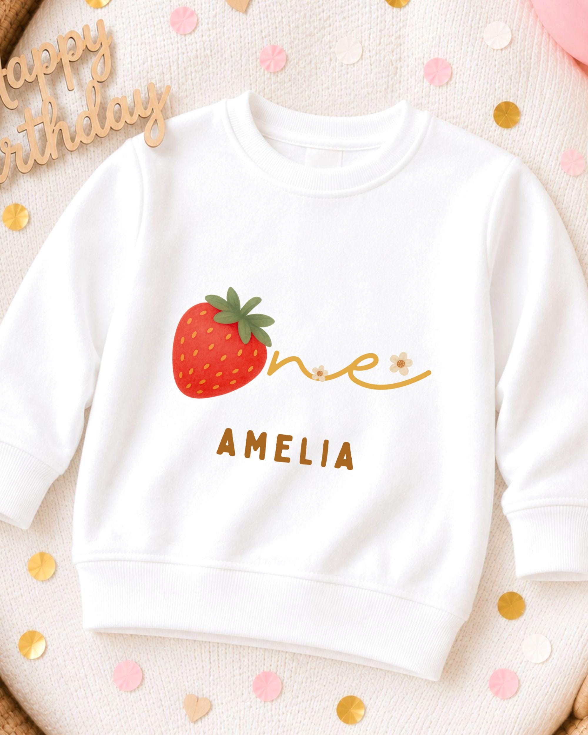 A white sweatshirt with a red strawberry and the name "Amelia" printed on it, along with confetti surrounding it.