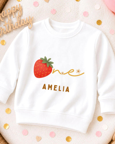 A white sweatshirt with a red strawberry and the name "Amelia" printed on it, along with confetti surrounding it.