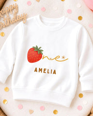 A white sweatshirt with a red strawberry and the name "Amelia" printed on it, along with confetti surrounding it.