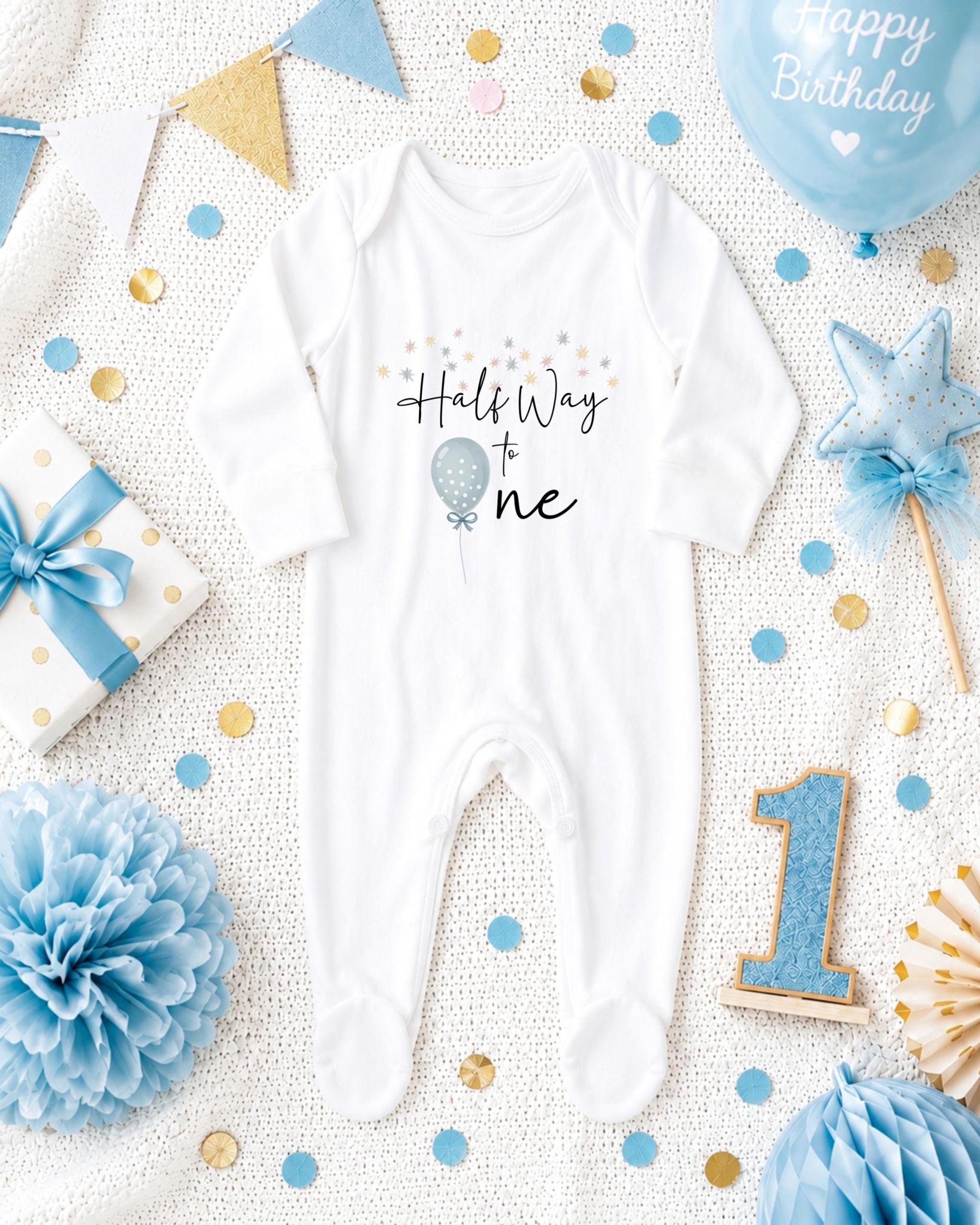 a baby onesie with the text "Half Way to One" printed on it, surrounded by various baby-themed items such as a blue balloon, a blue and gold polka dot gift box, a blue and gold star, a blue and gold confetti, a blue and gold pom pom, and a blue and gold number one.