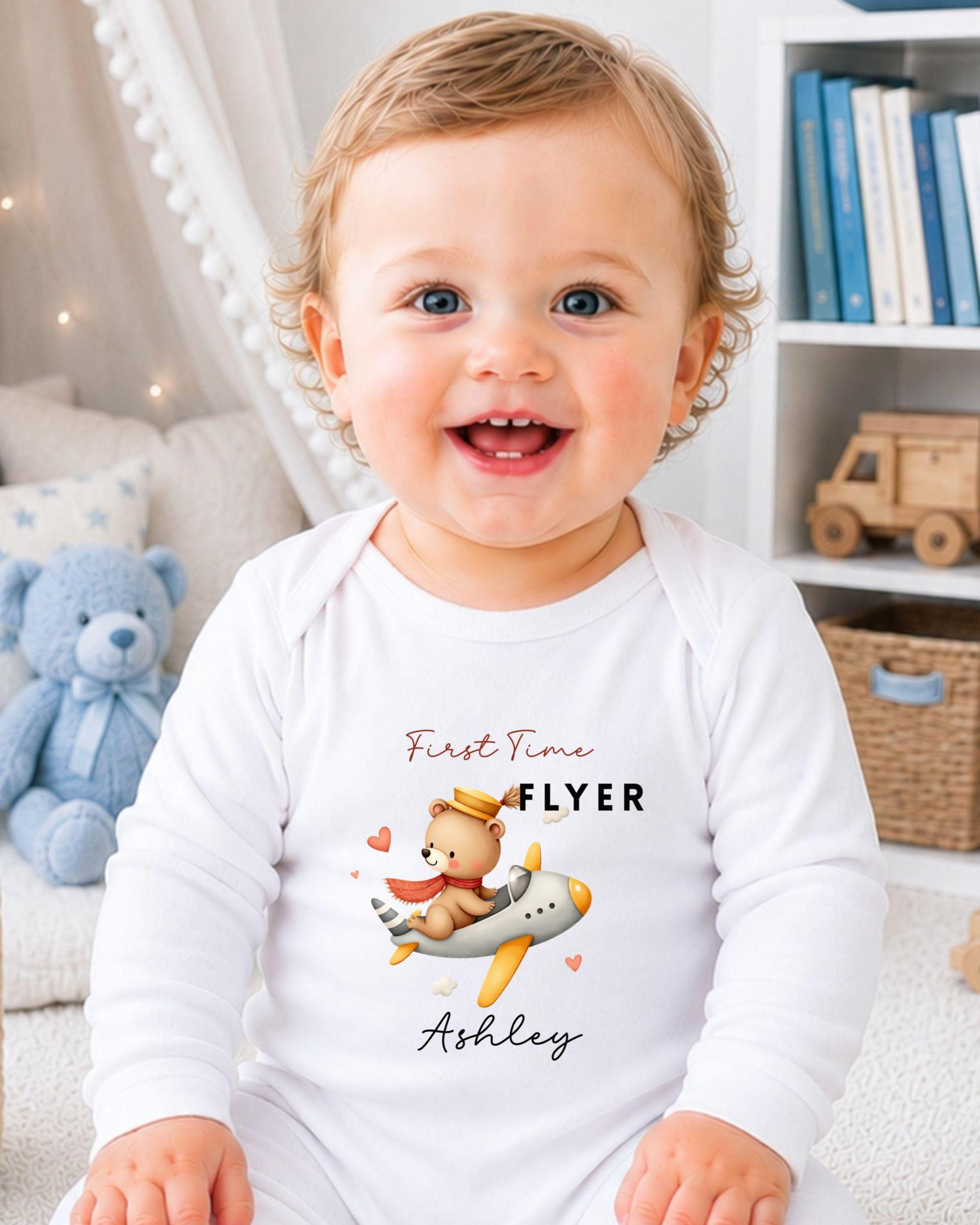 A smiling baby wearing a white onesie with a cartoon airplane and the text "First Time Flyer Ashley" printed on it.