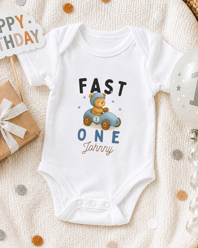 A white bodysuit with a cartoon image of a bear in a car and the text "FAST ONE Johnny" printed on it, along with a "Happy Birthday" sign and a balloon.