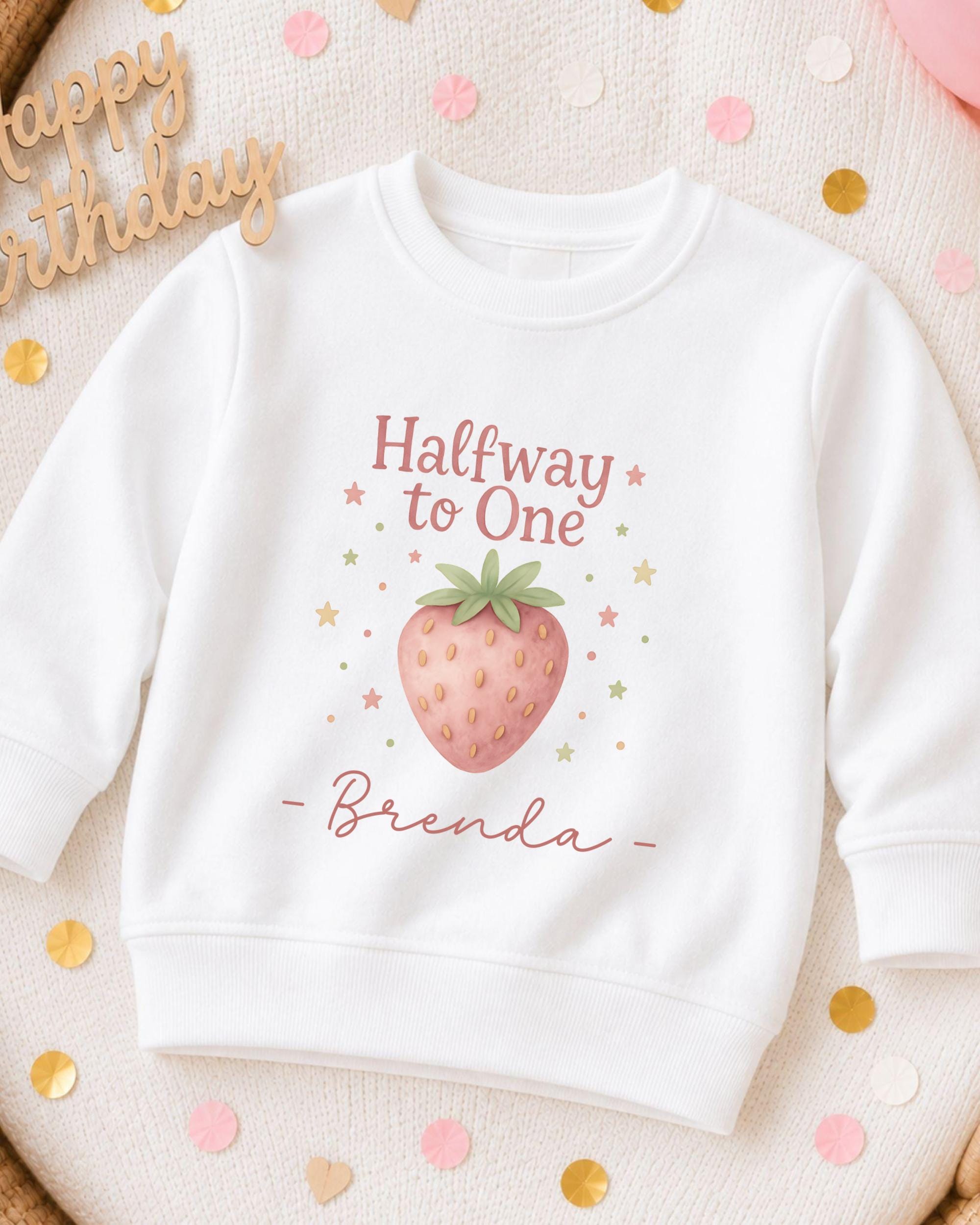A white sweatshirt with a strawberry graphic and the text "Halfway to One" printed on it, along with the name " Brenda" written below.