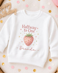 A white sweatshirt with a strawberry graphic and the text "Halfway to One" printed on it, along with the name " Brenda" written below.