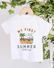 A white t-shirt with a graphic design depicting a beach scene, including a palm tree, beach chair, and various summer-themed objects. The text on the t-shirt reads "MY FIRST SUMMER Johnny".