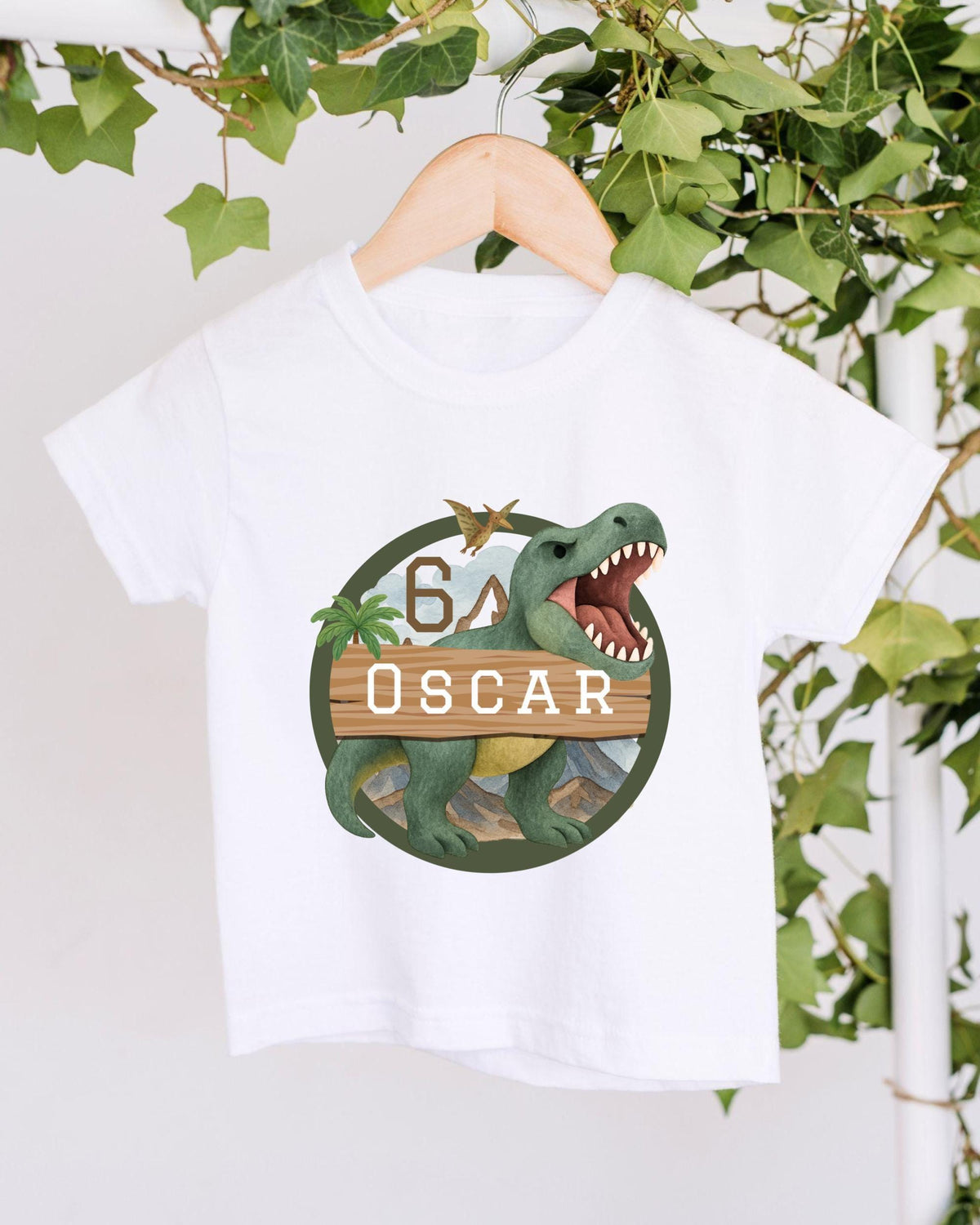 A white t-shirt with a graphic design featuring a dinosaur and the text "OSCAR" on it, hanging on a wooden hanger.
