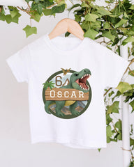 A white t-shirt with a graphic design featuring a dinosaur and the text "OSCAR" on it, hanging on a wooden hanger.