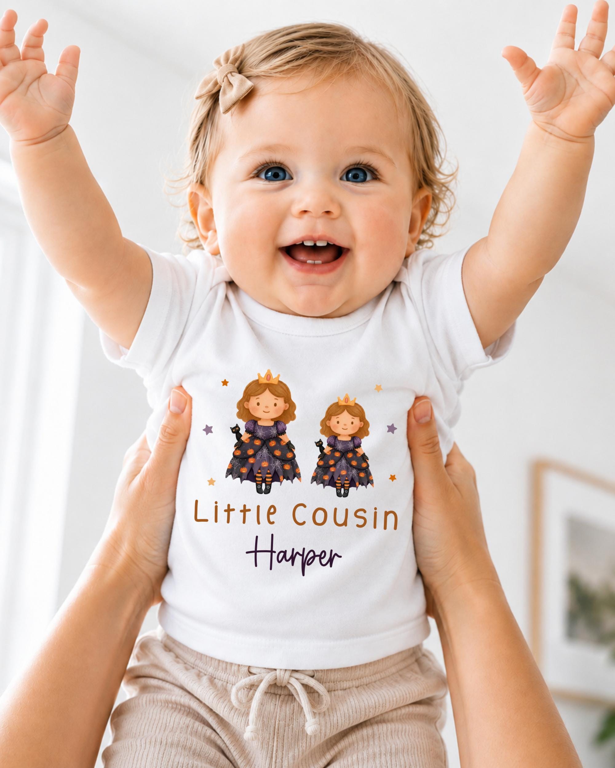 A smiling baby girl wearing a white t-shirt with a cartoon image of two princesses and the text "Little Cousin Harper" printed on it.