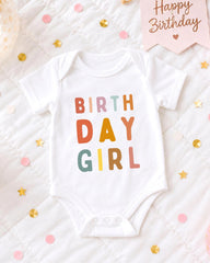 A white onesie with the text "BIRTHDAY GIRL" printed on it, surrounded by colorful confetti and a "Happy Birthday" sign.