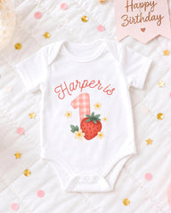 A white onesie with a strawberry and flower design, along with the text "Harper is" and the number "1", is displayed on a bed with confetti.